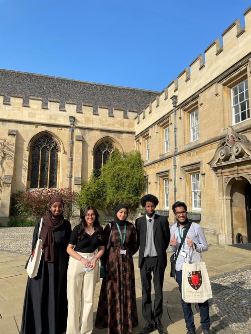 Five secondary school pupils stand in the St John's front quad on a sunny day smiling. One of them holds a tote bag with the St John's logo on it and does a thumbs up to the camera